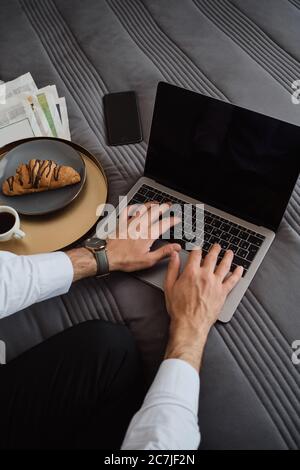 Nahaufnahme eines Mannes, der mit dem Laptop, Handy, Zeitungen und Croissant mit Kaffee auf dem Bett im modernen Hotel arbeitet Stockfoto