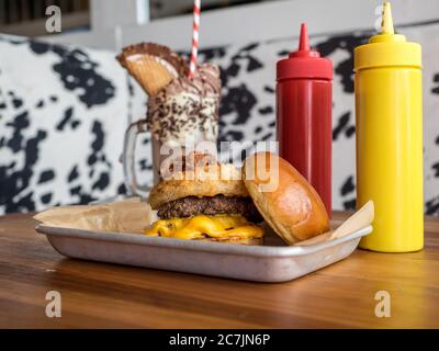 Burger auf einem Tablett mit Ketchup und Senf in der Nähe von A Schokoladenmilchshake auf einem Holztisch Stockfoto