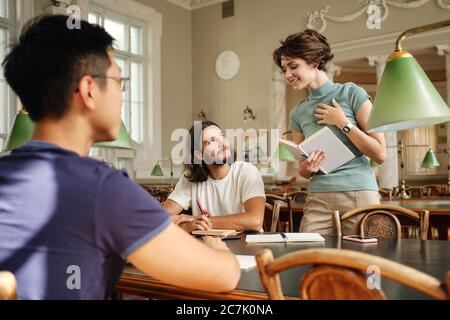 Junge attraktive lächelnde weibliche Tutor mit Buch glücklich erklärt den Studenten neues Thema während der Lektion in der Bibliothek der Universität Stockfoto