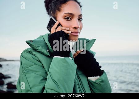 Portrait von attraktiven afroamerikanischen Mädchen in stilvollen Daunenjacke selbstbewusst in der Kamera im Gespräch auf Handy am Meer Stockfoto