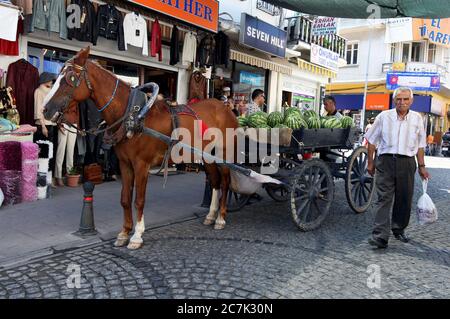 Ein türkischer Mann geht an einem Pferd und einem Wagen vorbei, der mit Wassermelonen beladen ist und auf einer Straße in Ayvalik in der Türkei geparkt ist. Stockfoto