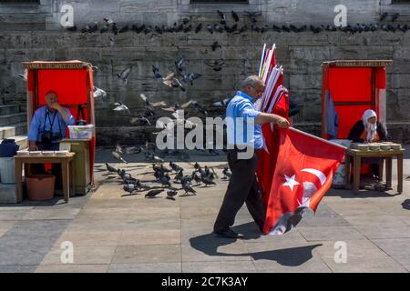 Ein türkischer Flaggenverkäufer kommt an Yeni Camii in Eminonu in Istanbul vorbei. In den Kabinen werden Getreideplatten verkauft, um die Tauben zu füttern. Stockfoto