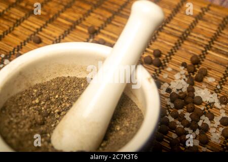 Gemahlenes schwarzes Piment in einem Keramikmörtel und schwarzes Piment und grobes Salz auf einer Schilfmatte verstreut. Nahaufnahme Stockfoto