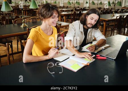 Zwei junge fröhliche Studenten, die sich beim gemeinsamen Lernen in der Bibliothek der Universität gerne Notizen machen Stockfoto