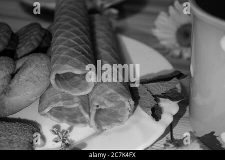 Waffelbrötchen und Kekse auf einem weißen Teller und eine Tasse Tee. Waffeln auf der Seite. Komposition auf einem hölzernen Hintergrund. Sichtbare Waffelstruktur. Schwarz und Stockfoto