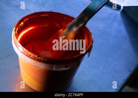 Die orange Farbe in der Dose steht auf einem Metalltisch und wird mit einem Holzspatel gerührt Stockfoto