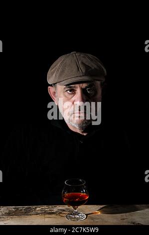 Ein älterer Mann mit einer Mütze sitzt an einem Tisch vor einem Glas Wein Stockfoto