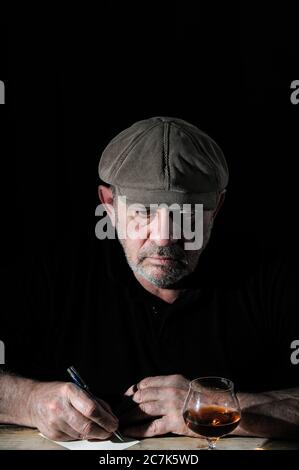 Ein älterer Mann mit Mütze schreibt auf einer Postkarte am Tisch Stockfoto