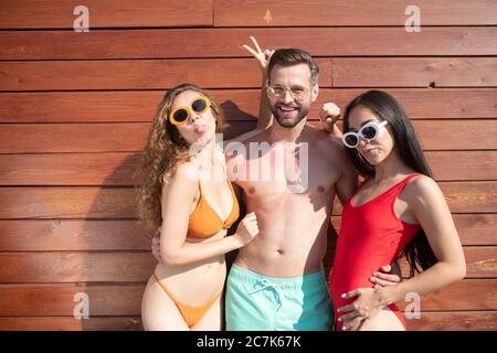 Junger, hübscher Mann, der zwei schöne Frauen in Schwimmanzügen umarmt Stockfoto