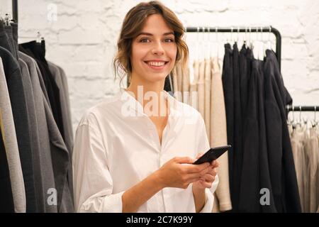 Junge schöne Casual Frau glücklich in der Kamera mit Handy in modernen Showroom suchen Stockfoto