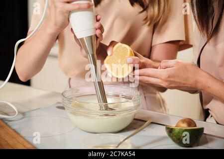 Hand der jungen Frau drückt frische Zitrone in Schüssel mit frischer Milch, während Teenager-Mädchen mit elektrischem Mixer mischen Eis Zutaten Stockfoto