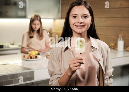 Brunette junge lächelnde Frau zeigt Ihnen hausgemachte eskimo Eis mit frischen Kiwi beim Essen vor der Kamera in der Küche Stockfoto