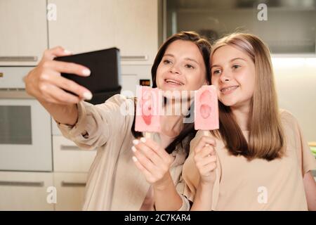 Zwei junge, fröhliche Weibchen mit langen Haaren machen Selfie in der Küche, während sie hausgemachtes eskimo-Eis mit Erdbeerscheiben essen Stockfoto