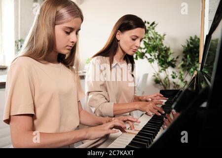 Serious blonde Teenager-Mädchen drücken Tasten des Klaviers während des Trainings während zu Hause Unterricht der Musik mit ihrer Mutter sitzt in der Nähe Stockfoto