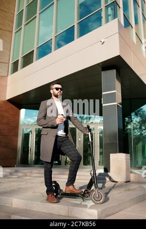 Serious junger Mann in Hosen, Mantel und Sonnenbrille auf Elektroroller durch moderne Architektur und mit einem Glas Kaffee stehen Stockfoto