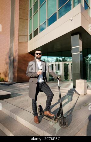 Junge elegante Mann in smart Casualwear mit Kaffee im Freien, während auf Elektroroller von moderner Architektur stehen Stockfoto