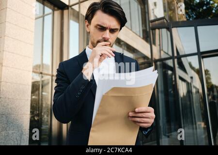Junge ernsthafte bärtige Brünette Mann in weißem Hemd und klassische Jacke halten Hand in der Nähe Mund im Freien stehend, während nachdenklich Dokumente lesen Stockfoto