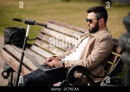 Junger Geschäftsmann mit mobilen Gadget Surfen im Netz, während auf Holzbank im Park an warmen Tag mit seinem Roller in der Nähe sitzen Stockfoto