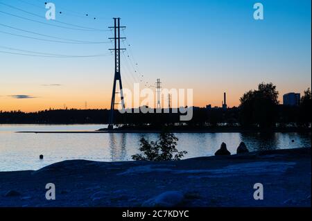 Zwei junge Frauen sitzen auf einer Klippe und beobachten die Silhouetten von hohen Pfosten gegen bunten Himmel während des Sonnenuntergangs. Stockfoto