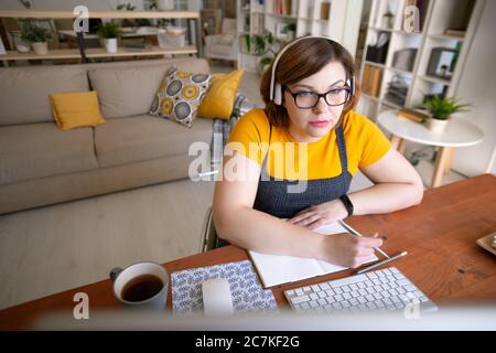 Moderne junge deaktivieren weibliche Schüler in Kopfhörer Blick auf Computer-Bildschirm und Notizen machen, während der Online-Kurs Stockfoto
