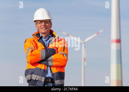 Lächelnder Ingenieur mit Hut und Schutzkleidung vor Windturbinen an einem sonnigen Tag - verschwommener Hintergrund Stockfoto