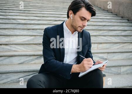 Junge attraktive bärtige Brünette Mann in weißem Hemd und klassischen Anzug mit drahtlosen Kopfhörer sitzen auf Treppen im Freien nachdenklich Notizen in Stockfoto