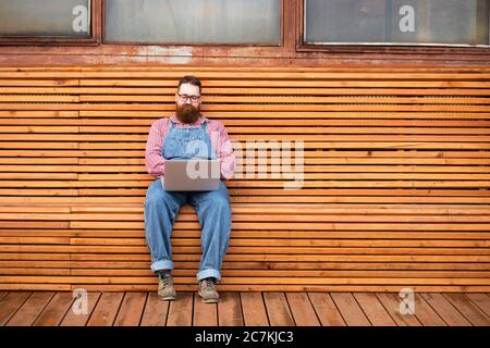 Porträt von einsamen brutalen bärtigen Hipster Mann in blauen Overalls, kariertes Hemd arbeiten auf Laptop sitzen auf Bank im Freien. Speicherplatz kopieren. Distanzauftrag Stockfoto