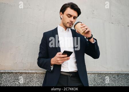 Junge gut aussehende bärtige Brünette Mann in weißem Hemd und klassischen Anzug mit drahtlosen Ohrhörern nachdenklich mit Handy, während Kaffee zu gehen trinken Stockfoto