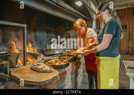 Zwei Frauen in der Nähe des Feuers Brot backen und traditionelle Zubereitung Slowenisches Essen aus Schweinegrippen Stockfoto