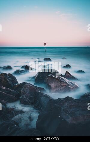 Wellen krachen über Felsen am Meer mit einem Strandmarker im Meer in der Ferne Stockfoto