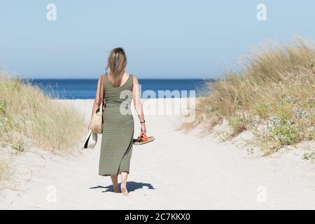 Eine blonde Frau mittleren Alters im Kleid, die barfuß auf einem sandigen Weg zum Meer mit ihren Sandalen in der einen Hand und einem Hut in der anderen geht. Stockfoto