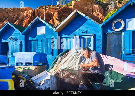 Europa, Portugal, Algarve, Litoral, Barlavento, Faro, in der Nähe von Albufeira, Praia dos Olhos de Agua, Fischer, die Netze ausbessern, blaue Fischerhütten vor den rotbraunen Klippen Stockfoto