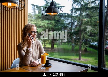 Junge charmante Frau mit Mobiltelefon, während allein im Coffee Shop in der freien Zeit, attraktiven weiblichen mit niedlichen Lächeln in Sprechen Stockfoto