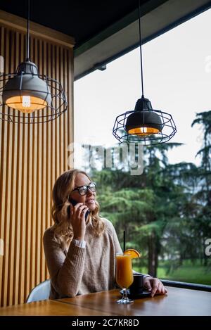 Junge charmante Frau mit Mobiltelefon, während allein im Coffee Shop in der freien Zeit, attraktiven weiblichen mit niedlichen Lächeln in Sprechen Stockfoto