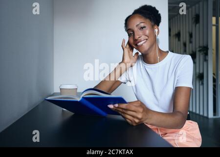 Ziemlich Casual afroamerikanische Mädchen in Kopfhörer mit Buch und Kaffee freudig in der Kamera in Co-Working-Raum suchen Stockfoto