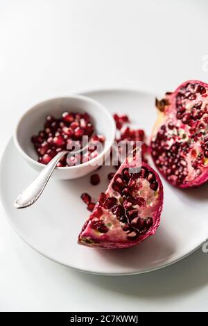 Rote Granatapfel in Scheiben geschnitten auf weißem Teller und weißem Hintergrund, lose Kerne in weißer Schale mit silbernem Löffel Stockfoto