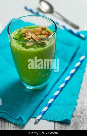 Grüner Smoothie in einem Glas auf einem türkisfarbenen Tuch mit zwei blauen und weißen Strohhalmen und einem silbernen Löffel Stockfoto