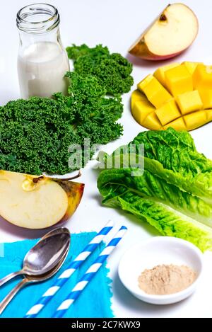 Alle Zutaten auf weißem Hintergrund verteilt, Milchflasche, Grünkohl, Mango-Igel, Apfelviertel, Romainsalat, zwei Strohhalme, zwei Silberlöffel, Nahrungsergänzungsmittel in einer weißen Schüssel Stockfoto