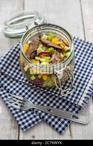 Rindsalat, buntes Gemüse, in einem Einmachglas auf weißem Holzhintergrund und blauem und weißem Karomuster, mit einer Campinggabel Stockfoto