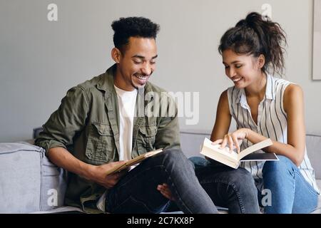 Junger lächelnder afroamerikanischer Mann und hübsche asiatische Frau lesen freudig Buch zusammen im Co-Working-Raum Stockfoto