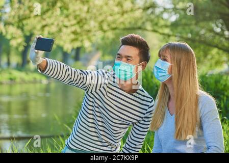 Mann und Frau tragen Gesichtsmaske sitzen auf einer Wiese, nehmen Sie ein Selfie, Corona Krise, Donau, Regensburg, Bayern, Deutschland Stockfoto