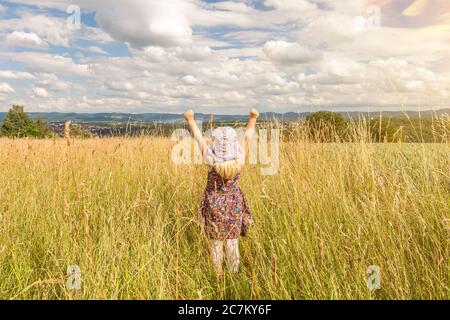 Nettes kleines blondes Mädchen, das im Sommer auf einem Feld steht und ihre Arme voller Freude hebt Stockfoto