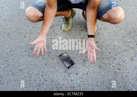 Guy ließ sein Handy auf den Asphalt fallen und brach es. Ein frustrierter Mann mit einem defekten Handy. Mann sitzt und hält ein defektes Smartphone Stockfoto