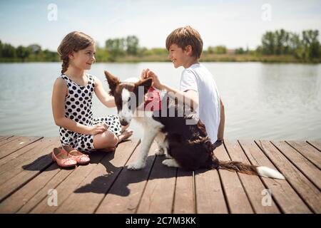 Kleine Kinder mit Hund sitzen zusammen auf Holz in der Nähe Teich und genießen. Stockfoto