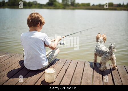 Lächelnder Junge, der Spaß am Fischen mit seinem Hund in einem Teich hat.Sommerfreude im Urlaub. Stockfoto