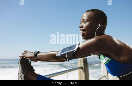 Lächelnde afrikanische Frau macht Bein Stretching Übung auf Geländer an der Küstenstraße. Fitness-Frau beim Aufwärmen auf der Straße am Meer. Stockfoto