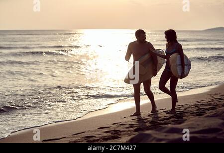 Junges Paar, das am Sandstrand bei Sonnenuntergang spazierengeht, Surfbretter trägt, redet Stockfoto