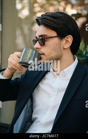 Junge attraktive bärtige Brünette Mann in weißem Hemd, klassische Jacke und Sonnenbrille verträumt neben beim Kaffee trinken auf der Straße der Stadt Stockfoto