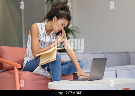 Junge ernsthafte beiläufige asiatische Frau im Gespräch auf Handy selbstbewusst auf Laptop arbeiten in Co-Working-Raum Stockfoto