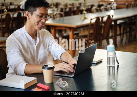 Junge lächelnde asiatische Student in Brillen glücklich arbeiten an Studie Projekt auf Laptop in der Bibliothek der Universität Stockfoto